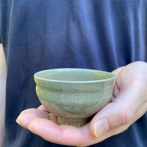 May include: A small, handmade ceramic bowl with a light green glaze. The bowl has a rounded shape and a short, pedestal base. The exterior has a textured appearance. The bowl is held in an open hand.