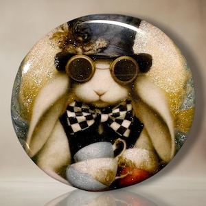May include: A circular decorative plaque featuring a white rabbit wearing a black top hat with gold accents, dark round goggles, and a black and white checkered bow tie. The rabbit holds a light blue teacup, with a glittery gold and white background.