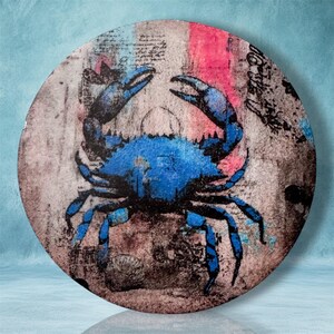May include: A circular wall art piece featuring a vibrant blue crab illustration centered on a distressed, textured background with faded script, black ink splatters, and vertical streaks of pink and grey tones across the surface.