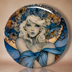 May include: A circular decorative plate featuring a detailed illustration of a person with wavy blonde hair, wearing a blue garment. The background displays a large golden crescent moon surrounded by clusters of blue and orange floral patterns.