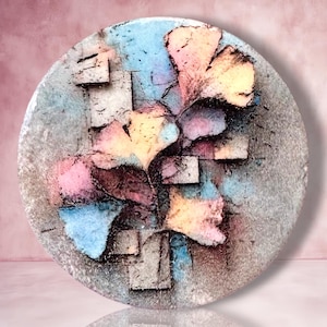 May include: A circular wall art piece featuring a textured, multi-layered composition of ginkgo leaf shapes and geometric square blocks. The surface displays a vibrant color palette of soft pink, light blue, and yellow, with dark accents.