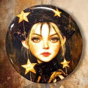 May include: A circular pinback button featuring a digital illustration of a person with blonde hair, freckles, and red lips. The person wears a dark hat adorned with glowing yellow stars, and additional yellow stars hang from the hat's brim.