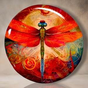 May include: A circular decorative plaque featuring a vibrant dragonfly with large blue eyes and orange wings. The body is green with intricate patterns, set against a background of swirling red, gold, and blue abstract shapes with a glossy finish.