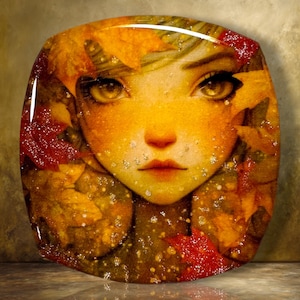 May include: A decorative glass plate featuring a detailed illustration of a face surrounded by vibrant autumn leaves in shades of red, orange, and gold. The surface has a glossy finish with small, scattered white specks resembling light reflections.