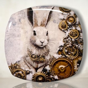 May include: A square decorative tile featuring a detailed illustration of a white rabbit with long ears. The rabbit is adorned with a necklace of metallic gold gears, surrounded by various brass-colored clockwork cogs and gears on a textured background.