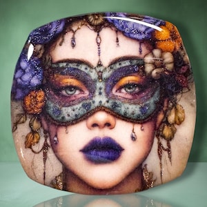 May include: A square decorative plate featuring a detailed illustration of a face wearing an ornate, glittery teal and purple masquerade mask, deep purple lipstick, and a crown of purple and orange flowers with dangling beaded accents.