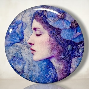 May include: A circular decorative plate featuring a profile illustration of a person with closed eyes, surrounded by large blue poppy flowers. The artwork uses a color palette of deep blues, purples, and soft skin tones with intricate line details.