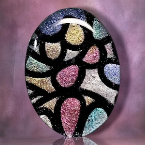 May include: An oval glass cabochon featuring a black grid pattern filled with various sections of sparkling glitter in shades of blue, gold, silver, pink, and teal. The surface is smooth and reflective, creating a multi-colored mosaic effect.