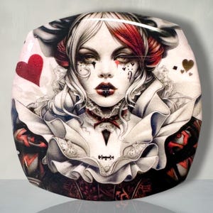 May include: A glossy, rounded decorative plaque featuring a detailed illustration of a person with pale skin, dark eye makeup, and red and white hair. The figure wears a ruffled white collar and dark clothing, accented by red heart symbols.