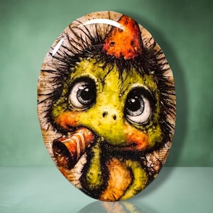 May include: An oval-shaped decorative plaque featuring a colorful illustration of a green, spiky creature with large eyes, wearing an orange party hat and holding a striped party blower. The background of the artwork incorporates vintage script text.