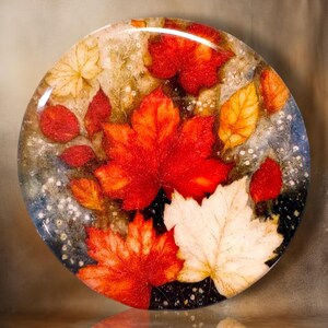 May include: A circular decorative plate featuring a vibrant autumn-themed design. The surface displays a collection of pressed maple leaves in shades of deep red, bright orange, golden yellow, and cream, accented by small white floral details.