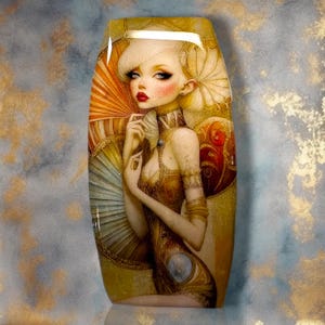 May include: A rectangular decorative plaque featuring a stylized illustration of a person with blonde hair, red lips, and pale skin. The figure is adorned in gold-toned attire and is positioned in front of large, layered, fan-like circular elements.