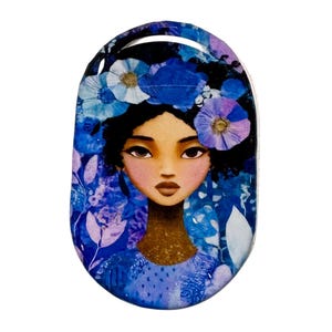 May include: Oval-shaped art piece featuring a woman with dark hair adorned with blue and purple floral accents. The woman's face is centered, with a blue floral background. The art piece has a handle at the top.