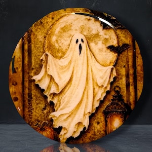 May include: A circular decorative plate featuring a central white ghost figure against a large, textured yellow moon. The background includes dark silhouettes of flying bats, a glowing lantern on the right, and a pumpkin at the bottom left.