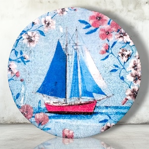 May include: A circular decorative plate featuring a sailboat with bright blue sails and a pink hull. The scene is set against a light blue textured background, framed by delicate illustrations of pink and white cherry blossom flowers.