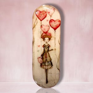 May include: An elongated oval decorative tray featuring a whimsical illustration of a person with brown hair wearing a dark dress, holding strings attached to three red heart-shaped balloons and one smaller pale pink heart-shaped balloon.