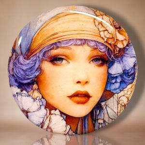 May include: A circular decorative plaque featuring a detailed illustration of a person with wavy purple hair, wearing a tan headband and surrounded by large blue and white flowers. The artwork has a glossy finish and a smooth, rounded edge.