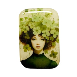 May include: A rectangular pendant featuring a woman with a floral headdress made of green clovers. The woman has brown hair and is wearing a green top. The background is a light beige color.