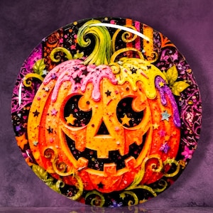 May include: A circular decorative plaque featuring a carved orange pumpkin with a jack-o'-lantern face. The pumpkin is adorned with dripping pink and yellow icing-like textures, small black and colored star shapes, and swirling green vines.