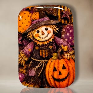 May include: A rectangular decorative plaque featuring a colorful illustration of a scarecrow wearing a purple hat and overalls, standing next to a large orange jack-o'-lantern. The background includes purple polka-dot patterns and orange shapes.