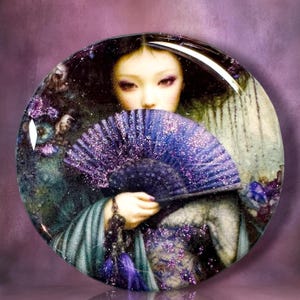 May include: A circular glass cabochon featuring a detailed illustration of a person with dark hair holding a vibrant purple glitter folding fan. The artwork displays deep blue and purple tones with sparkling accents, set against a dark background.