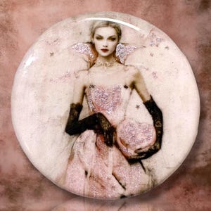 May include: A circular pin featuring a sepia-toned illustration of a person in a light pink dress with a high collar, wearing long black lace gloves and holding a pink heart-shaped object. The design includes shimmering glitter accents.