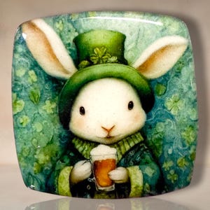May include: A square decorative tile featuring a white rabbit wearing a green top hat adorned with a shamrock. The rabbit is dressed in a green coat and holds a glass of amber-colored beer with white foam against a green background with clover patterns.