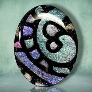 May include: An oval-shaped glass cabochon featuring a black base with embedded iridescent glitter fragments in shades of teal, purple, silver, and gold. The surface is smooth and reflective, displaying a mosaic-like pattern of shimmering colors.