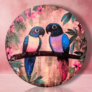 May include: A circular wall plaque featuring a printed illustration of two parrots perched on a branch. The parrots have blue heads, dark eyes, and colorful bodies in shades of pink, purple, and blue. The background shows pink flowers and green leaves.