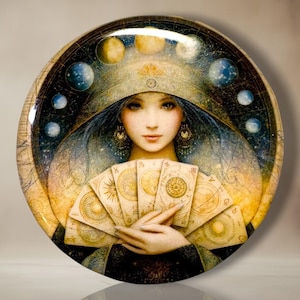 May include: A circular decorative plate featuring a central illustration of a figure holding a fan of six tarot cards. The background displays a celestial theme with golden suns, moons, and planetary spheres arranged in an arc around the figure.