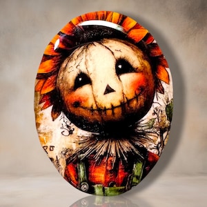 May include: An oval-shaped decorative piece featuring a stylized scarecrow head with a stitched mouth, large dark eyes, and a sunflower-like collar. The scarecrow wears a red and orange plaid shirt with green overalls, set against a textured background.