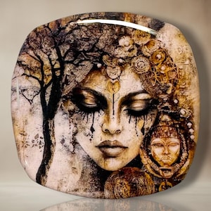 May include: A square decorative plate featuring a detailed illustration of a face with closed eyes, dark flowing makeup, and a tree silhouette. The artwork incorporates warm brown, gold, and black tones with intricate circular patterns and textures.