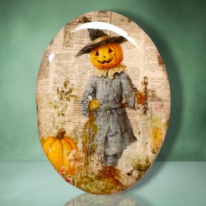 May include: An oval-shaped decorative plaque featuring a vintage-style illustration of a pumpkin-headed scarecrow wearing a dark hat and blue coat. The background consists of aged newspaper print, with a large orange pumpkin at the base.