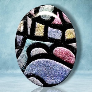 May include: An oval-shaped glass cabochon featuring a stained-glass style pattern with thick black lines separating segments of shimmering, glittery colors including blue, pink, silver, and gold. The surface has a smooth, glossy, and reflective finish.
