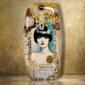 May include: A rectangular resin plaque featuring a central black and white portrait of a person with a bob haircut. The piece is embellished with gold glitter, white pearls, a fan-shaped element, and a vibrant turquoise accent surrounding the portrait.