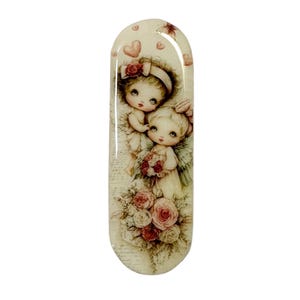 May include: An oval-shaped decorative item featuring a vintage illustration of two cherubic figures with wings, holding a bouquet of roses. The artwork has a soft, pastel color palette with pink and cream tones, and is embellished with hearts.