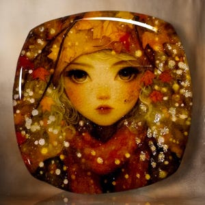 May include: A square glass plate featuring a detailed illustration of a person with large eyes wearing a hood adorned with orange and red autumn leaves. The design includes scattered white and yellow circular accents resembling falling snow or light.