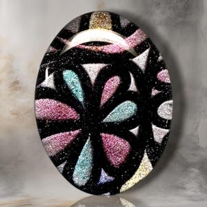 May include: An oval-shaped cabochon featuring a black background with a repeating floral-inspired pattern. The design consists of glittery, textured petals in shades of pink, teal, silver, and gold, separated by bold, smooth black lines.