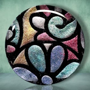 May include: A circular decorative object featuring a stained glass-style design with thick black outlines separating segments of shimmering, glittery colors including gold, silver, teal, pink, and purple, all under a smooth, glossy finish.