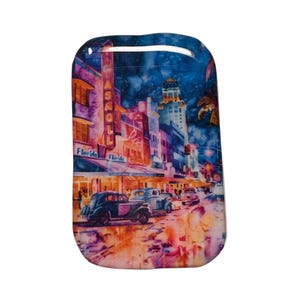 May include: A rectangular, rounded-top keychain featuring a watercolor painting of a vintage Miami street scene. The artwork depicts classic cars, buildings, and the text "Florida". The color palette includes blues, oranges, and purples.