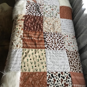 May include: A patchwork quilt featuring various western-themed patterns including cacti, horseshoes, cow print, and script text. The quilt includes white faux fur accents on the sides and a label indicating dimensions of 54 inches by 36 inches.