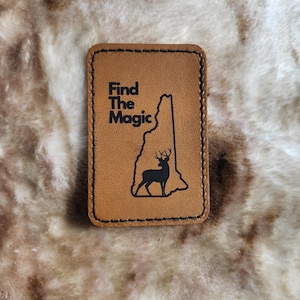 May include: A rectangular, tan leather patch with the words "Find The Magic" in black, along with a black silhouette of the state of New Hampshire and a deer. The patch has a stitched border.