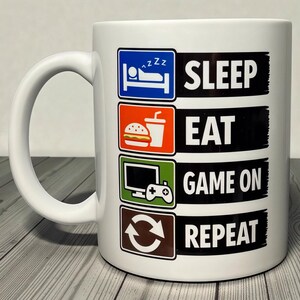 Może przedstawiać: Biały ceramiczny kubek z czarnym napisem "SLEEP EAT GAME ON REPEAT". Kubek posiada ikony łóżka, burgera i napoju, ekranu komputera z kontrolerem do gier oraz symbolu recyklingu. Kubek ma biały uchwyt.