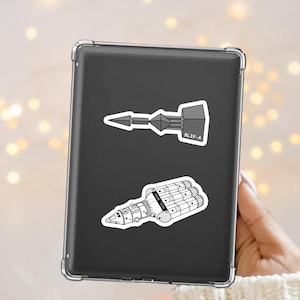 May include: Two black and white stickers on a tablet. One sticker features a geometric design with the text "BLIP-A". The other sticker depicts a detailed, stylized spaceship. The stickers are on a black tablet.