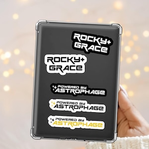 May include: A selection of stickers on a black tablet. The stickers display the text "ROCKY+GRACE" and "POWERED BY ASTROPHAGE" in different fonts and colours, including white, black, and yellow. The tablet is held by a person wearing a white jumper.