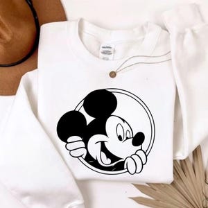 Pu&ograve; includere: Felpa bianca girocollo con grafica in bianco e nero di Topolino. Il disegno mostra il viso e le mani di Topolino che escono da un cerchio. Una collana color oro &egrave; visibile vicino al colletto.
