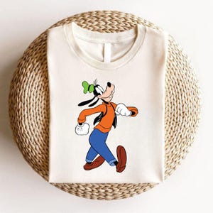 Puede incluir: Una camiseta blanca con una imagen de dibujos animados de Goofy caminando. Goofy lleva una camisa roja, pantalones azules y un sombrero verde.