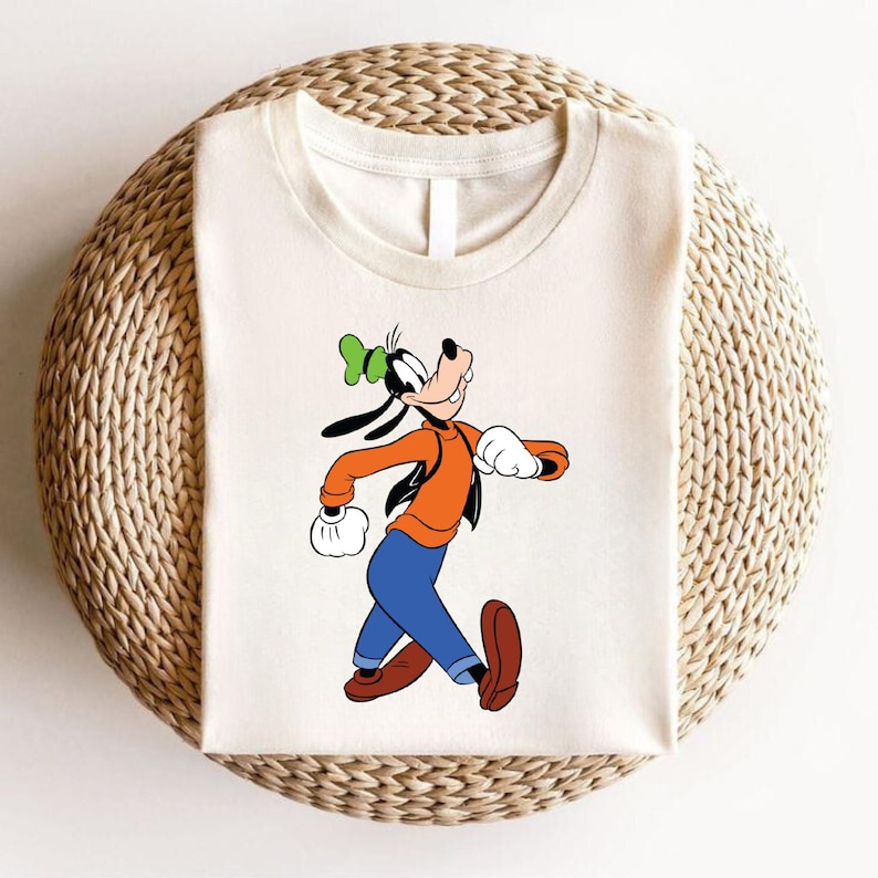 Puede incluir: Una camiseta blanca con una imagen de dibujos animados de Goofy caminando. Goofy lleva una camisa roja, pantalones azules y un sombrero verde.