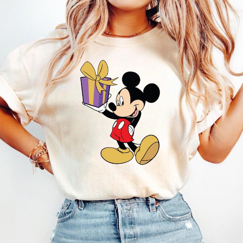 Puede incluir: Una camiseta blanca con una imagen de dibujos animados de Mickey Mouse sosteniendo una caja de regalo morada y amarilla.