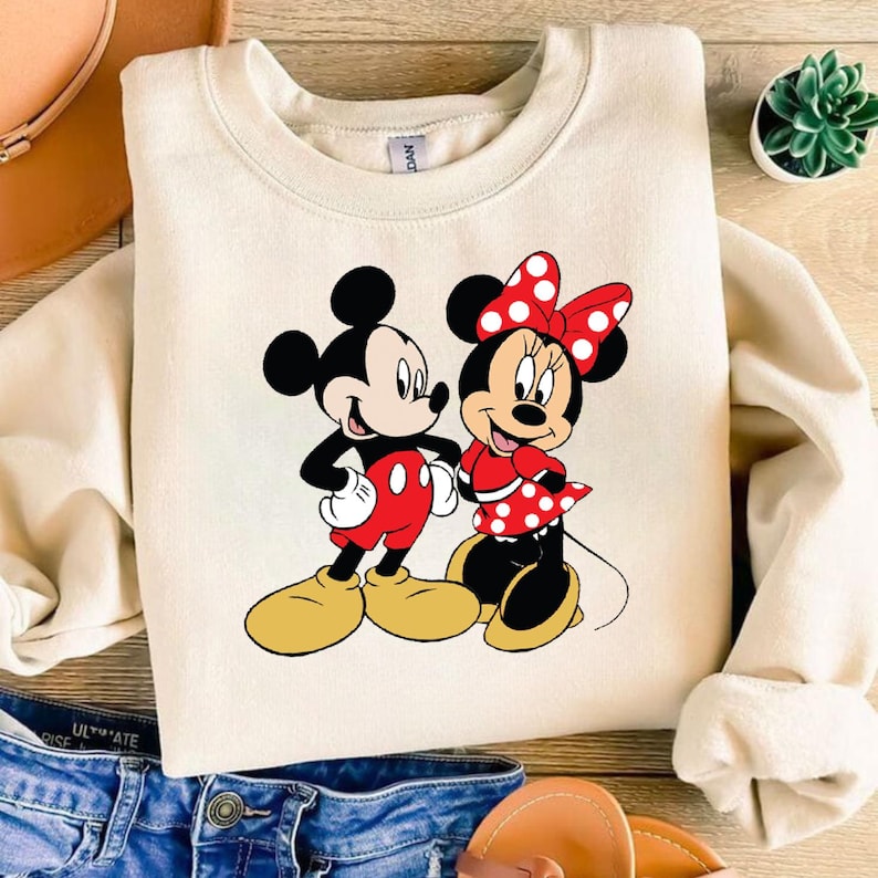 Puede incluir: Una sudadera de color crema con un gr&aacute;fico de Mickey Mouse y Minnie Mouse. Mickey lleva pantalones cortos rojos y Minnie lleva un vestido de lunares rojos y blancos.